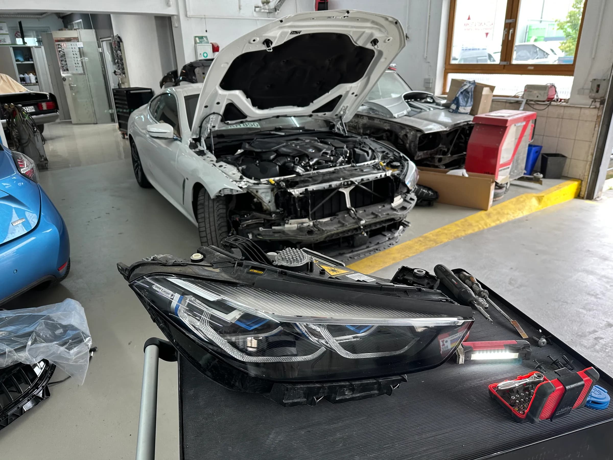 Weißer BMW mit zerlegter Front in der Werkstatt, der ausgebaute Scheinwerfer liegt im Vordergrund auf der Werkbank.