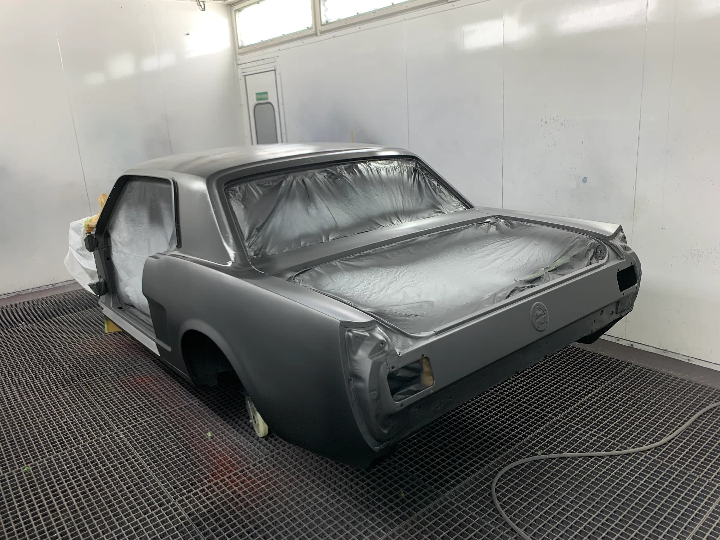 Karosserie eines Ford Mustang aus den 1960ern in grauer Grundierung in der Lackierkabine, die Scheibenausschnitte sind mit Folie abgeklebt.