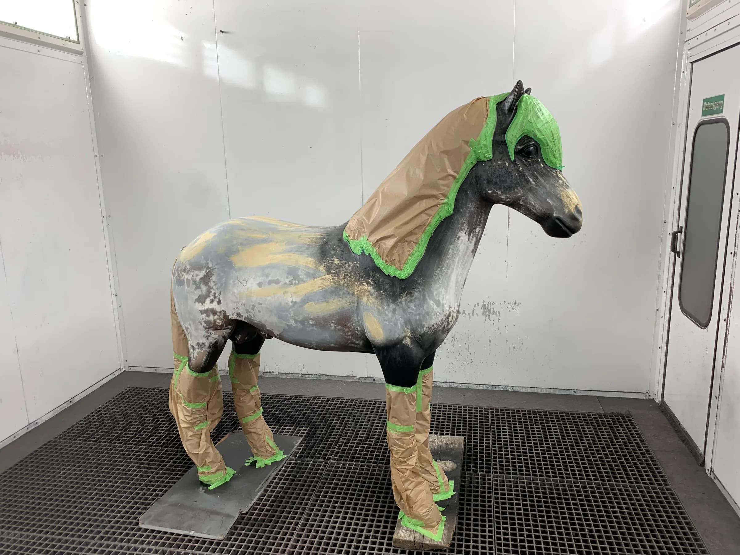 Lebensgroße Pferdefigur in der Lackierkabine, mit Abklebepapier und grünem Klebeband an Kopf, Mähne und Beinen maskiert.
